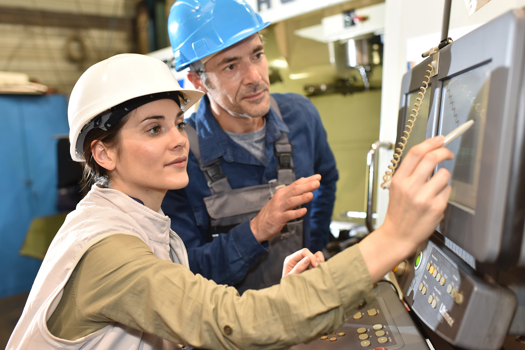 Woman and man wearing a hard hat on a factory floor. Woman is using a PC tablet that is connected to factory machines to input informaiton while man watches