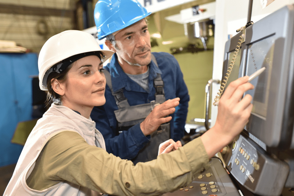 woman wearing a hard hat using a tablet device on a factory floor while being monitored by manager